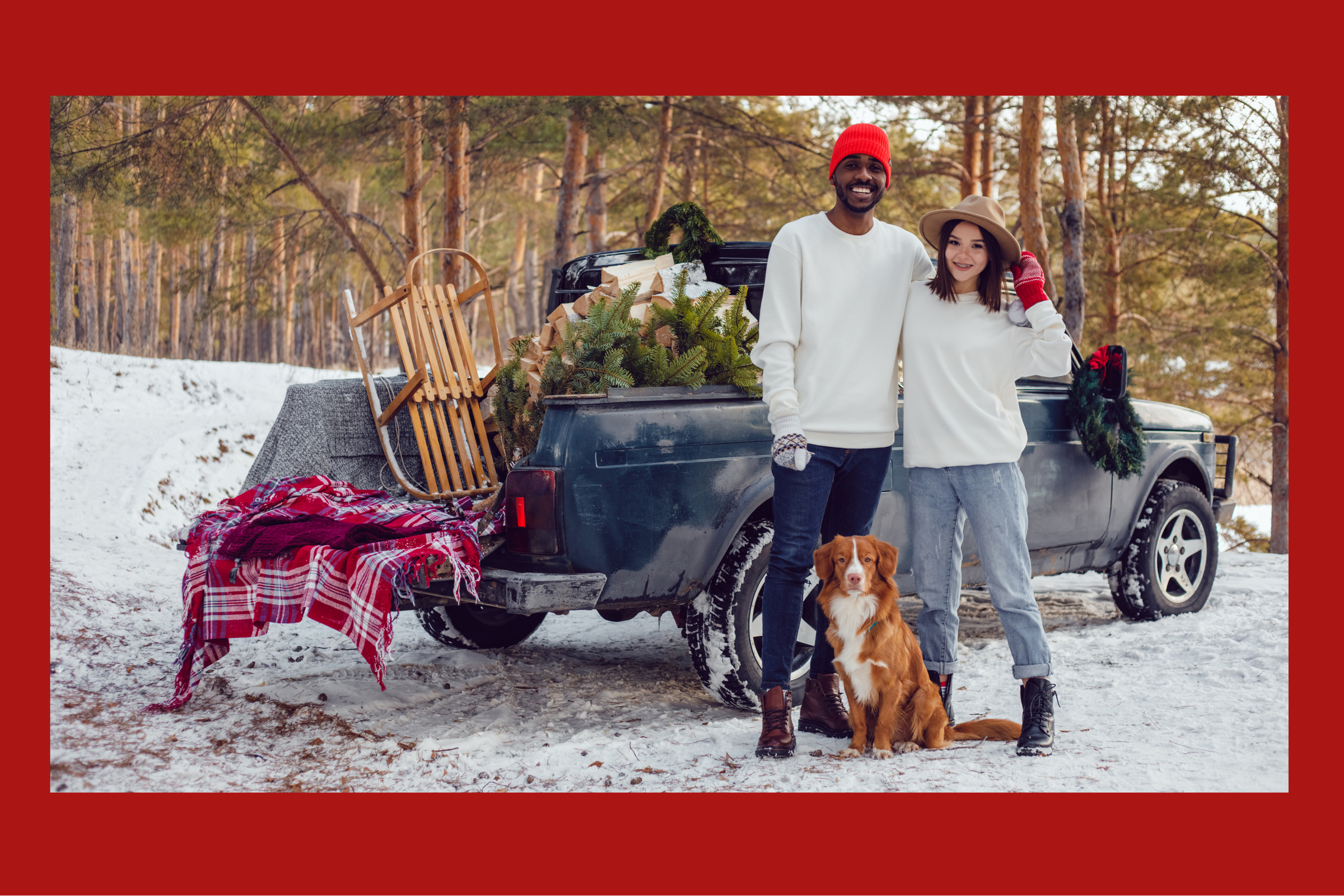 Couple with a dog standing in front of a truck loaded with Christmas decorations in a snowy setting.