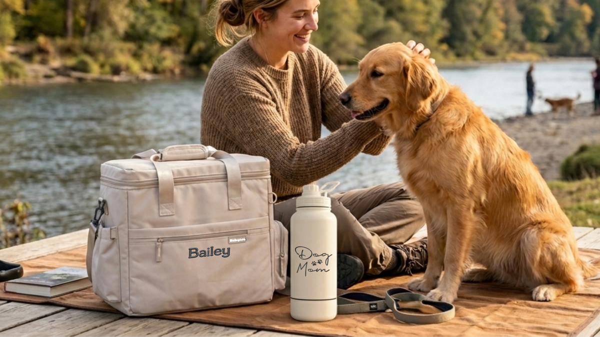 A woman sits on a wooden dock by a lake, petting a golden retriever. In front of them are outdoor gear items: a beige insulated bag labeled Bailey, a white water bottle with the words Dirty Paws, and a leash.